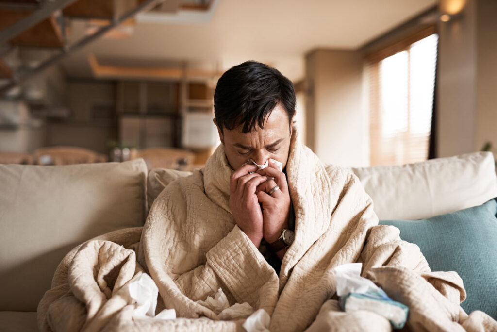 Hombre con gripe bajo una manta en el sofá sonándose la nariz